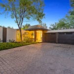 Modern house exterior with a brick driveway, two-car garage, large windows, and a flat roof. Surrounded by trees and greenery, with a tree in the front yard. Located at 23544 Highland Glen Dr in Santa Clarita.