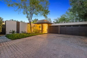 Modern house exterior with a brick driveway, two-car garage, large windows, and a flat roof. Surrounded by trees and greenery, with a tree in the front yard. Located at 23544 Highland Glen Dr in Santa Clarita.