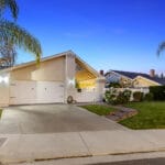 Single-story house with an illuminated exterior, two-car garage, and well-kept lawn, situated in the suburban Plaza Gavilan neighborhood of Santa Clarita, CA at twilight.