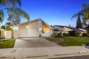 Single-story house with an illuminated exterior, two-car garage, and well-kept lawn, situated in the suburban Plaza Gavilan neighborhood of Santa Clarita, CA at twilight.