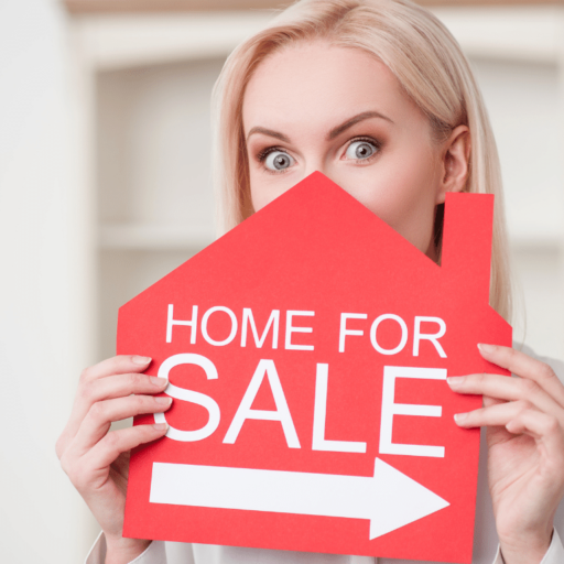 A person with blonde hair is holding a red house-shaped sign that reads "Home For Sale," partially covering their face.